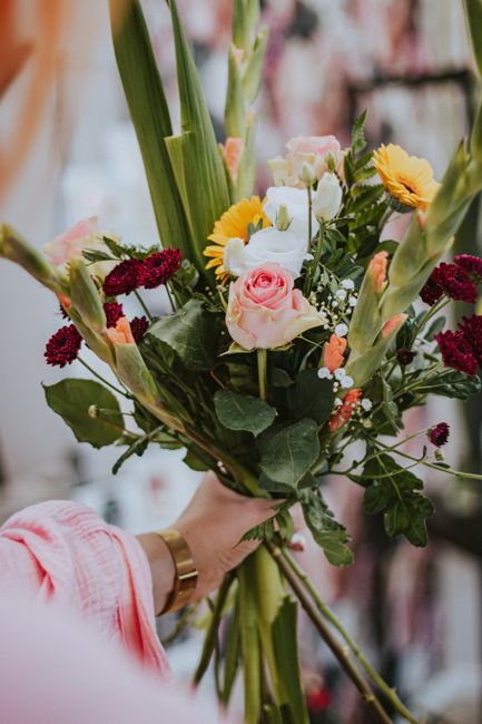 A person holding a vibrant floral bouquet featuring pink roses, white lisianthus, yellow and orange daisies, and deep red chrysanthemums, accented with lush green foliage and long, slender leaves. The