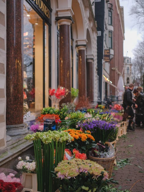 A vibrant outdoor floral display outside a shop on a city street, featuring a variety of fresh flowers arranged in baskets and pots. The arrangement includes white, orange, pink, purple, and green flo