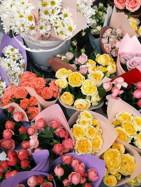 A vibrant display of fresh flower bouquets featuring a variety of blooms including white daisies with yellow centers, soft pink roses, bright yellow roses, and coral-colored roses. The flowers are arr